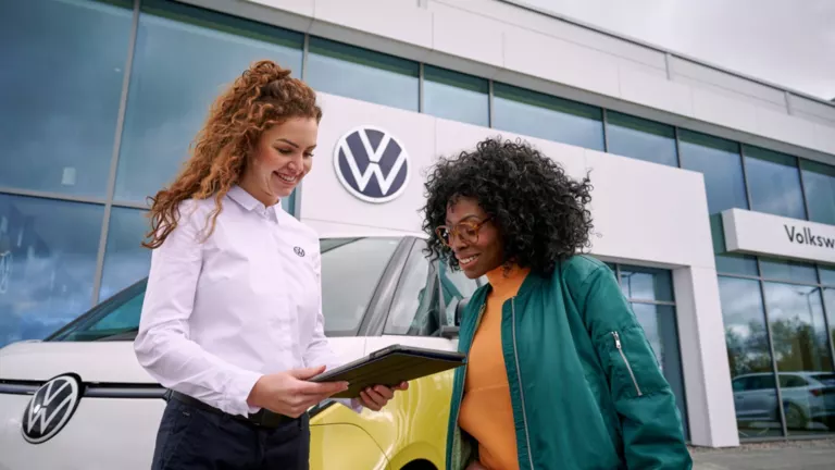 Eine VW-Mitarbeiterin steht mit einer Kundin vor einem VW-Autohaus und berät sie.