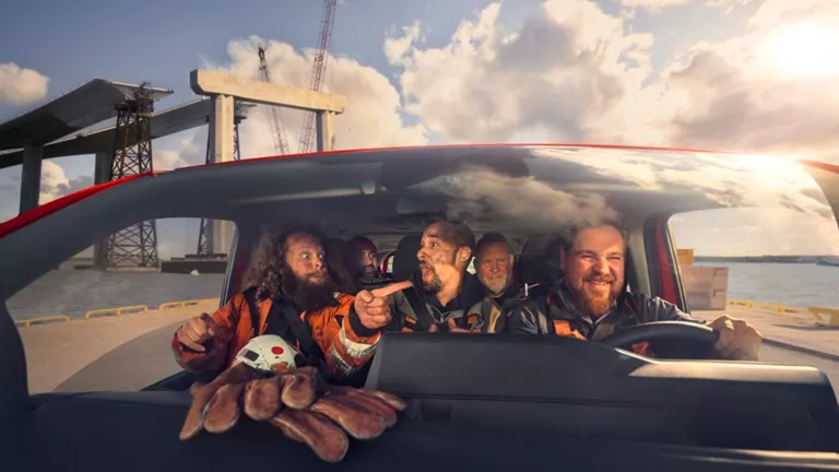 In einem VW Auto sitzen 5 gutgelaunte Männer in orangener Arbeitskleidung. Im Hintergrund ist der Berliner Alexanderplatz zu sehen.