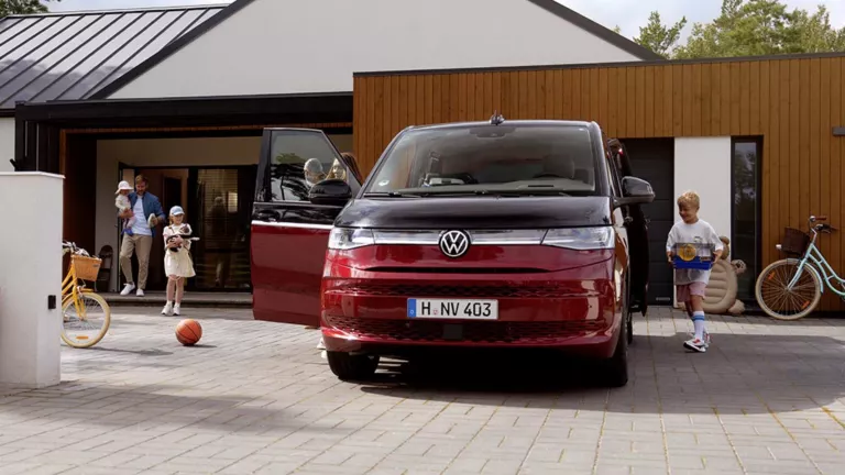 Eine vierköpfige Familie steht mit einem roten VW Multivan vor einem modernen Wohnhaus, wobei die Mutter und der ältere Junge das Auto beladen, während der Vater das Baby hält