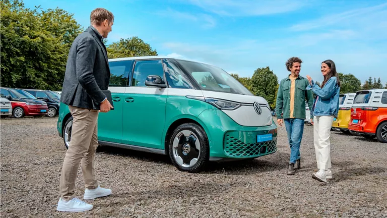 Eine Frau und ein Mann sehen sich einen Türkisen VW ID. Buzz auf einem Autohof an.