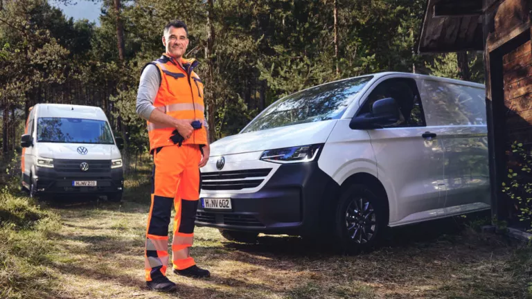 Arbeiter in Warnkleidung steht im Wald neben einem VW Transporter und einem VW Crafter vor einer Holzhütte