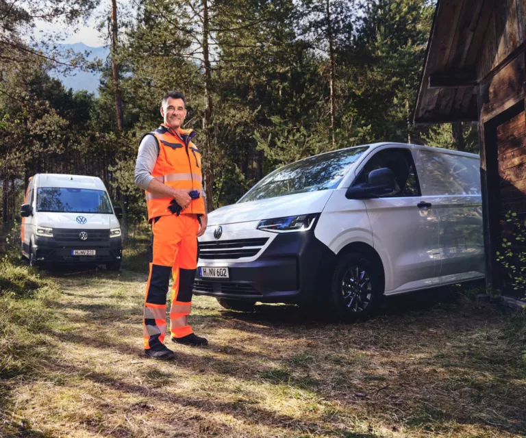 Arbeiter in Warnkleidung steht im Wald neben einem VW Transporter und einem VW Crafter vor einer Holzhütte