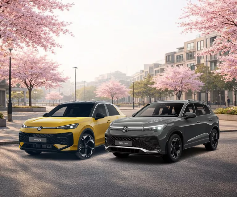 Ein grauer VW Tiguan und ein gelber VW T-Roc stehen auf einem asphaltiertem Weg in einer Stadt mit blühenden Bäumen.