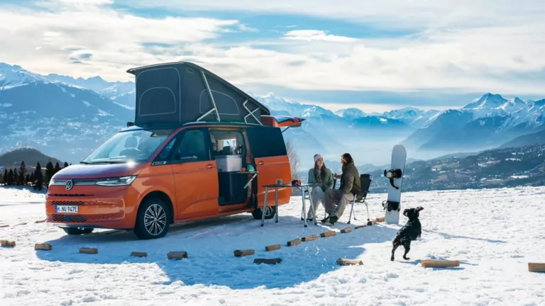 Ein VW California steht in einer Winterlandschaft in den Bergen. Zwei Menschen sitzen vor ihm auf Campingstühlen und unterhalten sich.