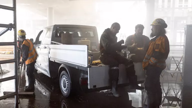 Ein weißer Transporter steht im Rohbau, auf seiner Pritsche frühstücken zwei Handwerker, daneben zwei Bauarbeiter.