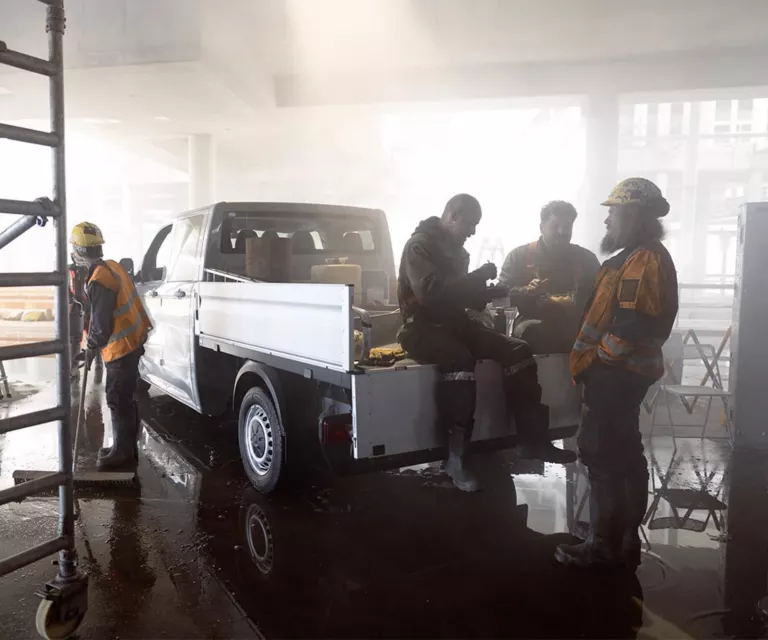 Ein weißer Transporter steht im Rohbau, auf seiner Pritsche frühstücken zwei Handwerker, daneben zwei Bauarbeiter.