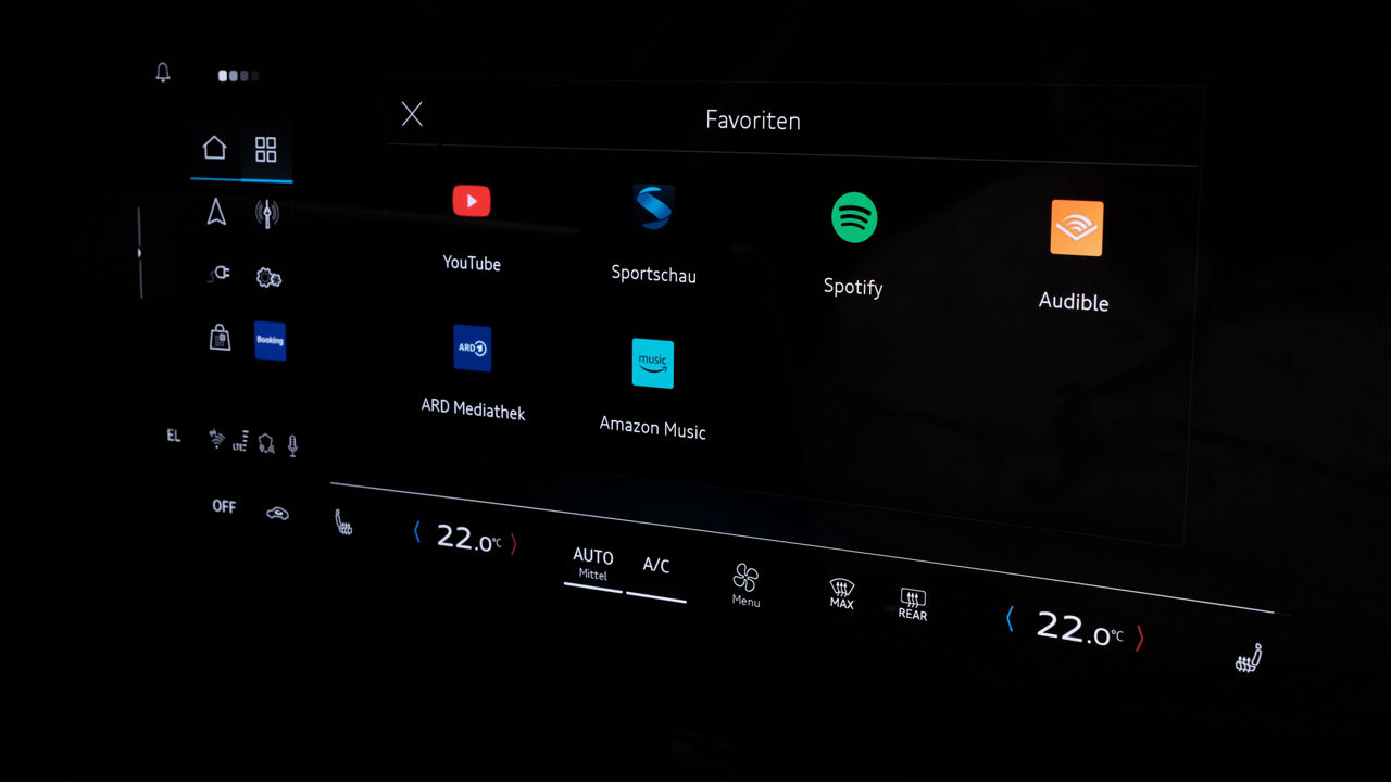 Favorites screen of a vehicle infotainment system showing a curated selection of media and entertainment apps. Visible app icons include YouTube, Spotify, Audible, Amazon Music, ARD Mediathek, and Sportschau. The central display is integrated into a dark cockpit with active climate controls, presenting entertainment applications as a core part of the connected in‑car digital ecosystem