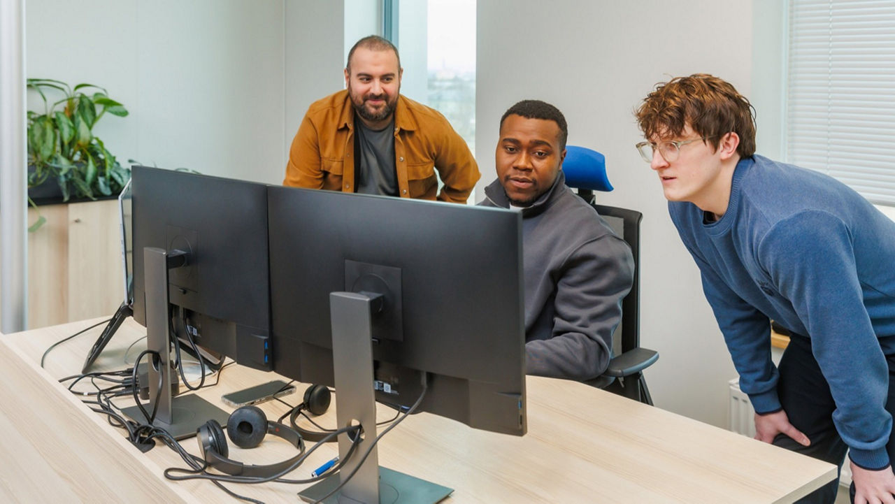 Three people at a desk with dual monitors in a bright office.