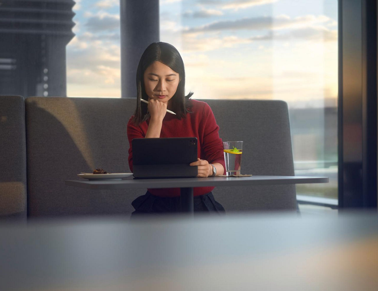 Young woman working on a tablet in a modern lounge area with large windows at sunset.
