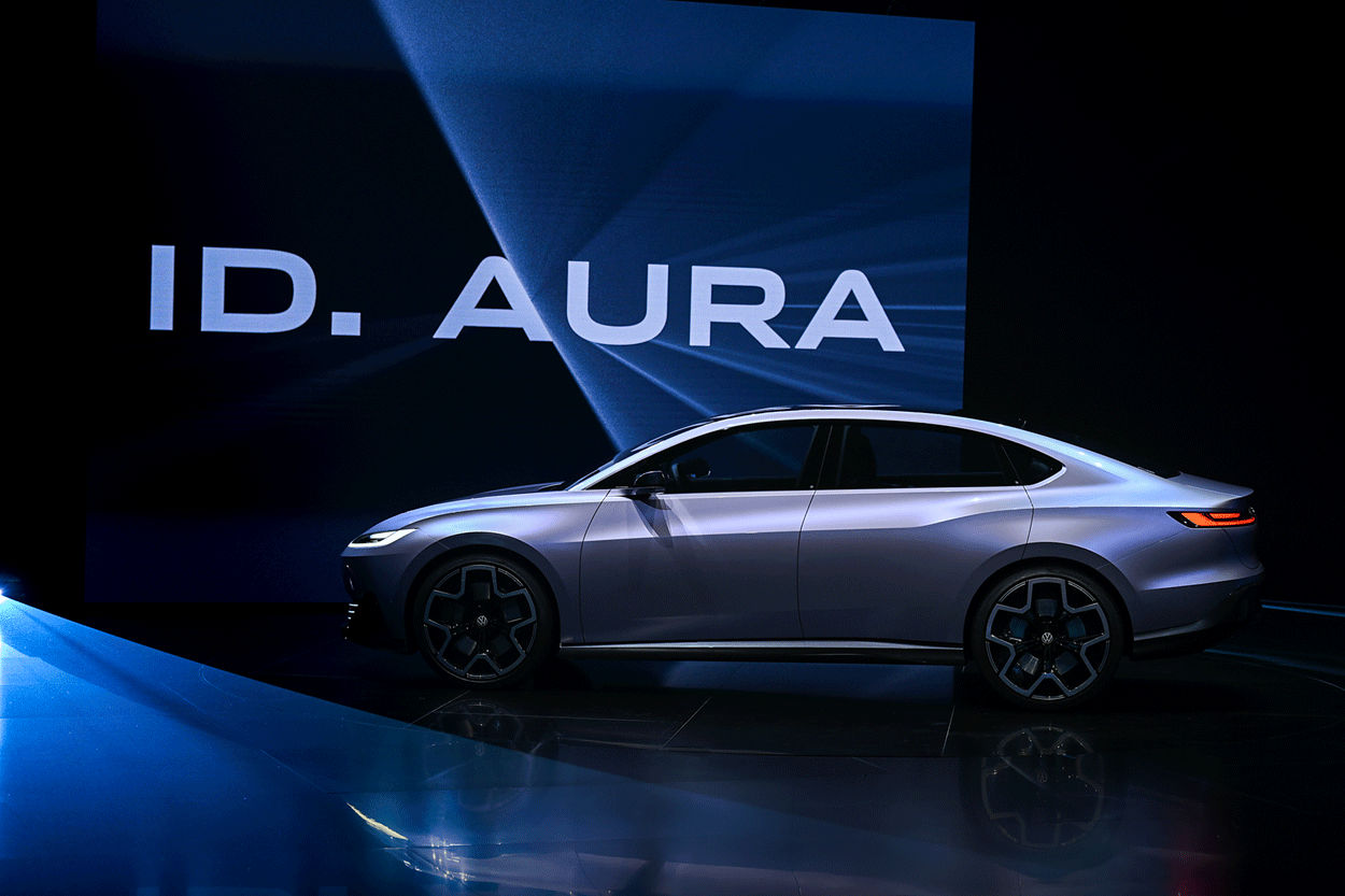 Side view of the ID. AURA on stage with a large screen in the background displaying 'ID. AURA'. Lighting highlights the car’s sleek design.
