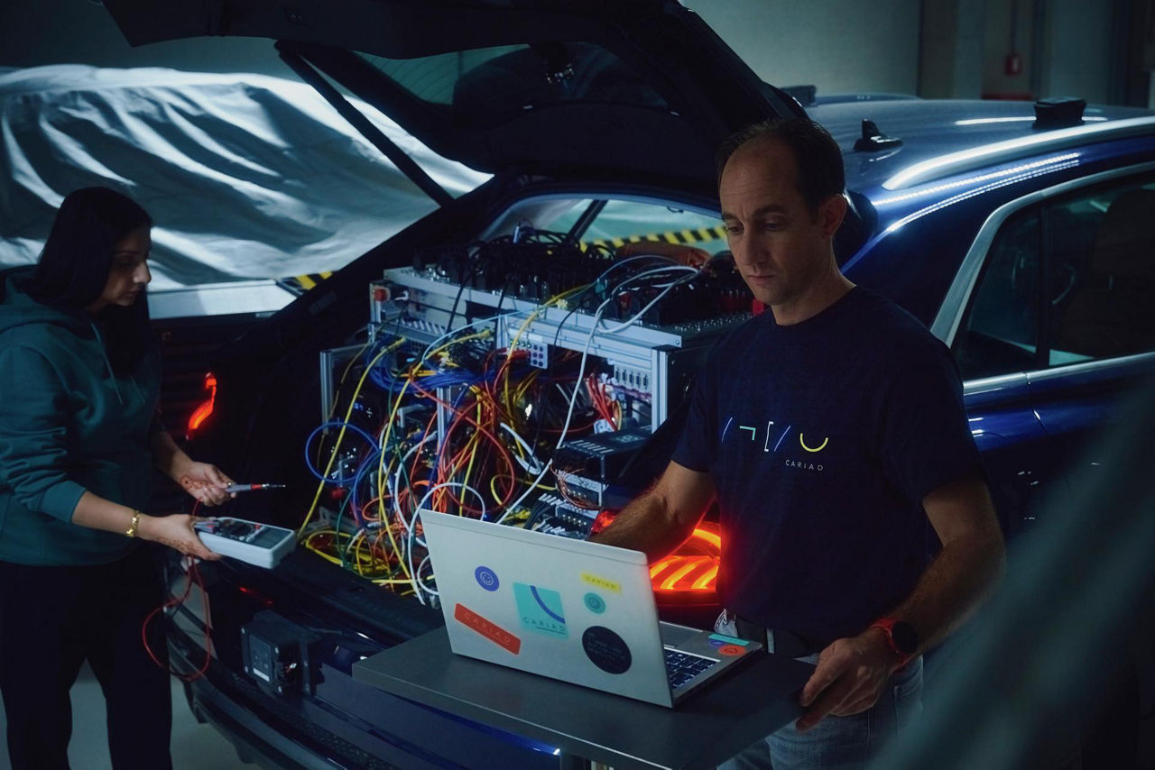 Two individuals working with cables and electronic equipment in the trunk of a car, one using a laptop.