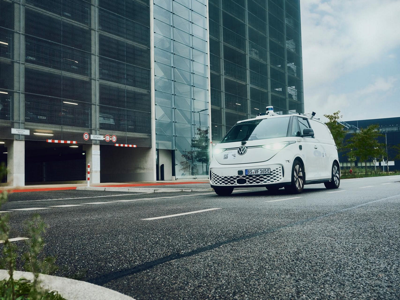A white Volkswagen van equipped with rooftop sensors navigates an urban environment, hinting at autonomous driving trials amid modern infrastructure.