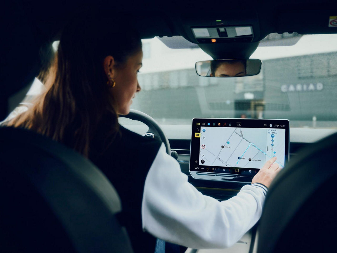 A driver engages with a touchscreen navigation system inside a car, parked near a building marked “CARIAD,” emphasizing connected mobility and tech integration.
