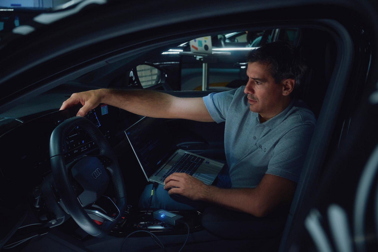 Person in driver's seat of car holding a laptop, performing diagnostic work with electronic devices and cables visible