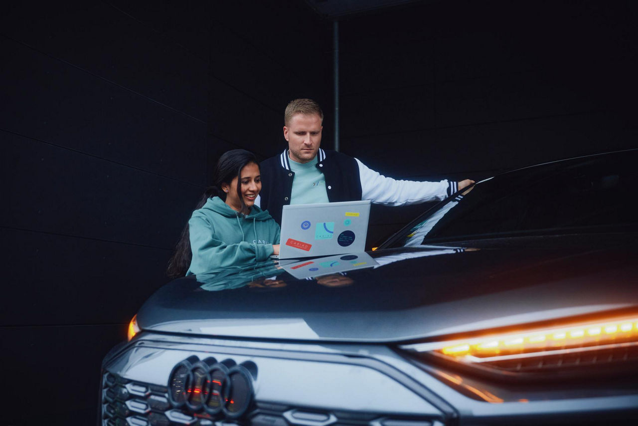 Two people standing by an Audi car with illuminated headlights, looking at a laptop on the car's hood.