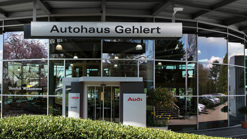 Eingang des Autohauses Gehlert mit Glasfassade, Schild 'Autohaus Gehlert' und Audi-Logo, umgeben von Büschen