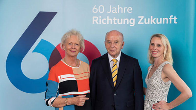 Drei Personen vor einer Wand mit dem Text '60 Jahre Richtung Zukunft', zwei Frauen und ein Mann, alle lächelnd und formell gekleidet