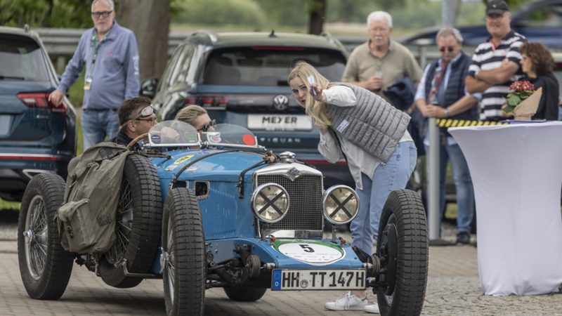 Ein blauer Oldtimer mit zwei Fahrern und eine Frau weist den Weg