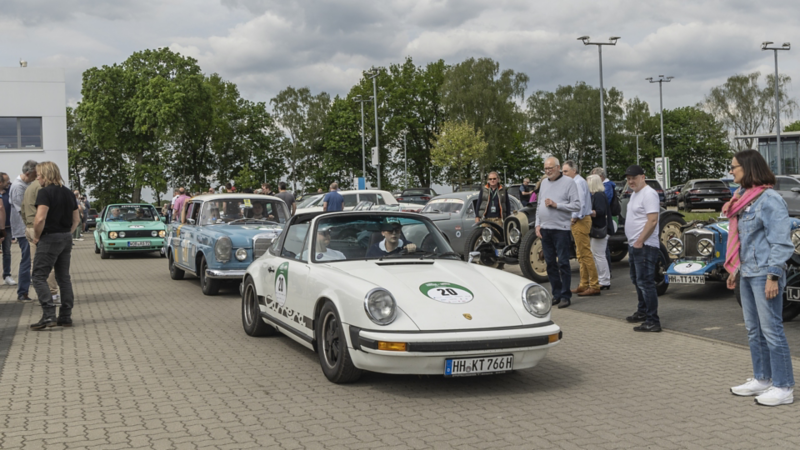 Außengelände mit Oldtimern und Menschen auf dem Parkplatz
