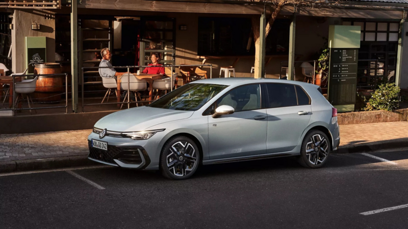 Silberner Volkswagen Golf geparkt vor einem Restaurant, zwei Personen sitzen draußen an einem Tisch