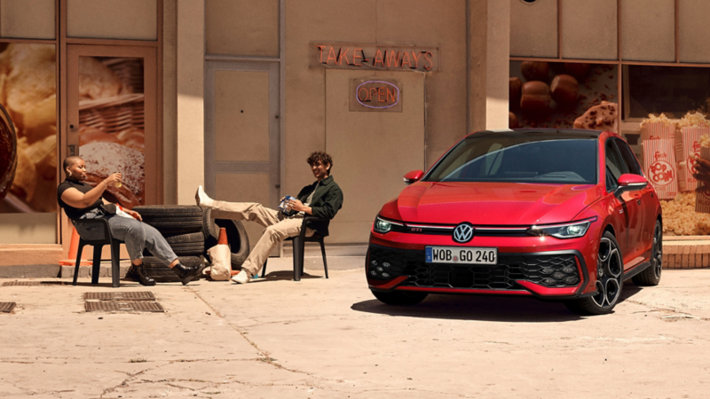 Ein roter VW Golf GTI steht vor einem Gebäude. Zwei Personen sitzen neben dem Wagen.