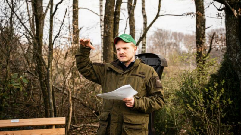Mann mit grüner Mütze und brauner Jacke hält Papiere und gestikuliert im Wald