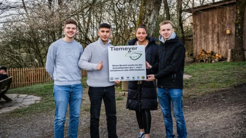 Vier junge Erwachsene stehen draußen und halten ein Schild mit der Aufschrift 'Tiemeyer' und Informationen zur Begrünung des Bereichs im Jahr 2023