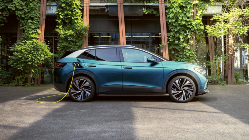 Seitenansicht eines blauen VW ID.4, das an ein gelbes Ladekabel angeschlossen ist. Das Auto steht vor einem Gebäude, dass mit grünen Pflanzen bewachsen ist. 