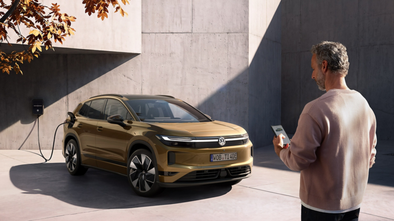 Ein Mann schaut auf das Handy in seiner Hand. Im Hintergrund lädt ein oliv-bronzefarbener VW ID. Tiguan an einer Wallbox.