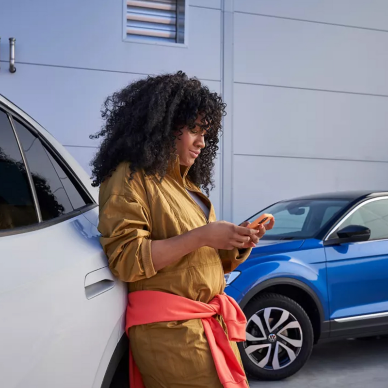 Μια κοπέλα ακουμπάει σε ένα VW και κοιτάει το myVolkswagen app στο κινητό της