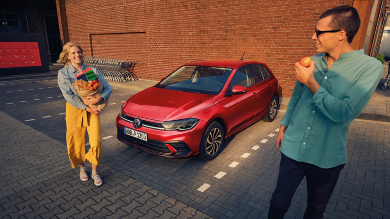 Ein roter VW Polo steht auf einem Parkplatz vor einem Supermarkt, ein lachendes Paar steht mit Einkäufen daneben.