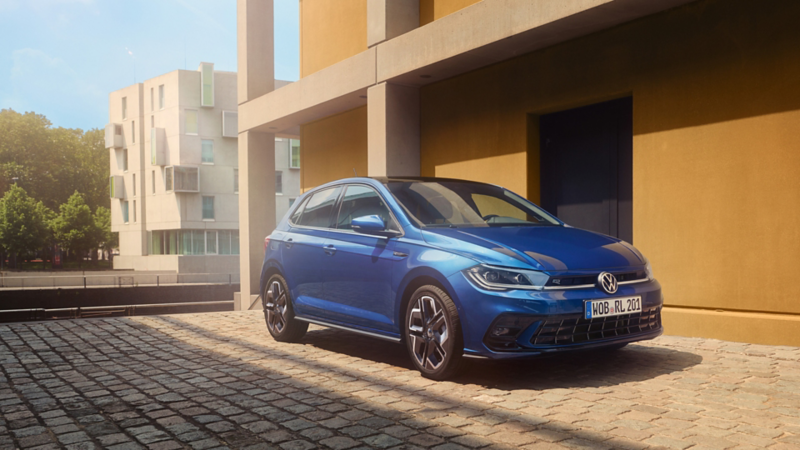Ein blauer VW Polo steht vor einem gelben Gebäude im Sonnenschein.