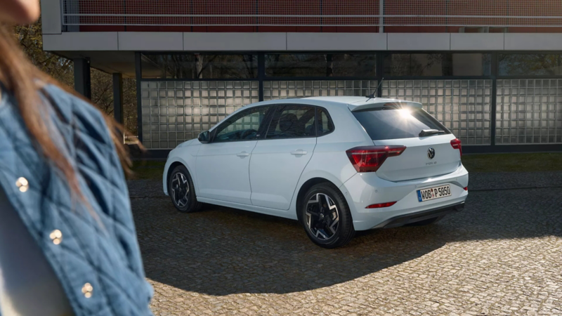 Ein hellblauer Polo parkt seitlich vor einem Gebäude und eine Frau steht vor ihm.