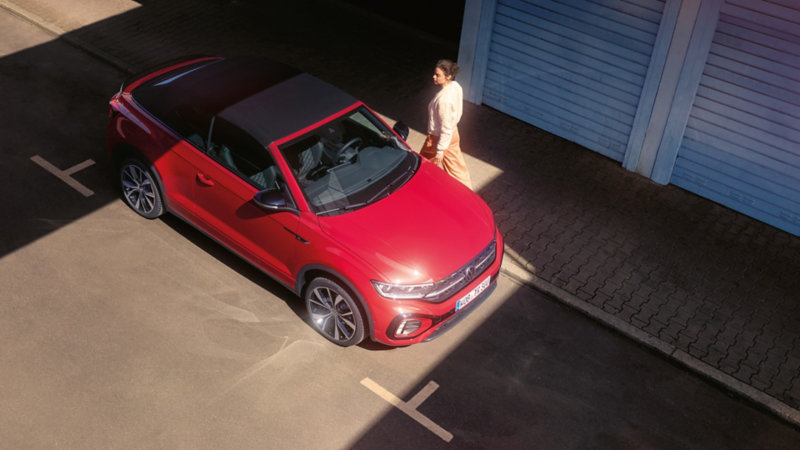 Vogelperspektive auf das rote VW T-Roc Cabriolet mit geöffnetem Dach. Parkend am Straßenrand, eine Frau geht vorbei. 