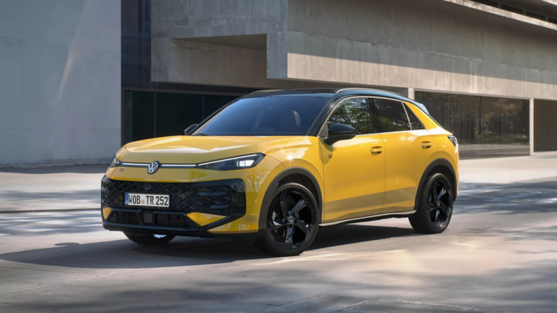 Seitenansicht eines gelben T-Roc R-Line der vor einem modernen Gebäude mit viel Beton steht. 