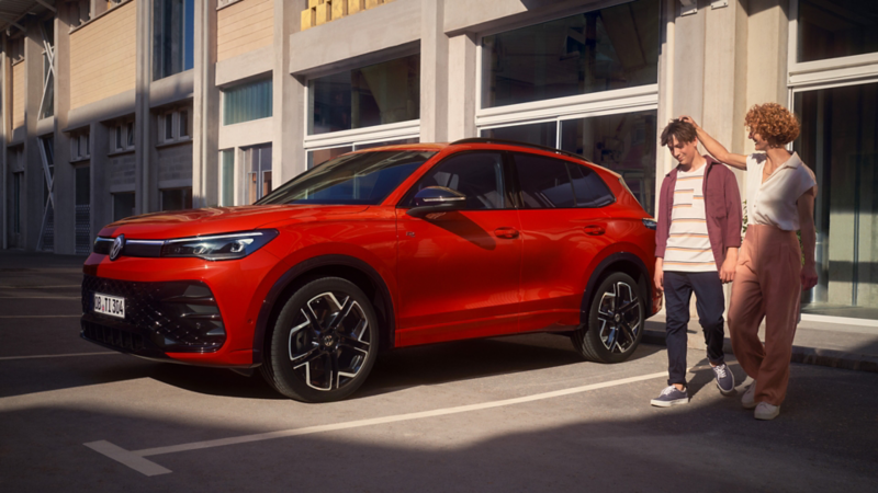 Eine Frau und ein Mann laufen auf der Straße vor einem parkenden roten VW Tiguan
