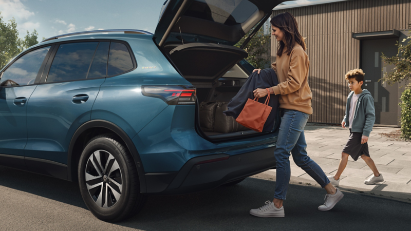 Frau lädt Gepäck in den geöffneten Kofferraum eines blauen VW Tiguan, Kind läuft daneben auf dem Bürgersteig