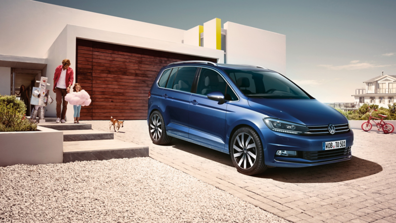 Blauer VW Touran steht vor einem modernen Einfamilienhaus, eine Familie geht darauf zu.