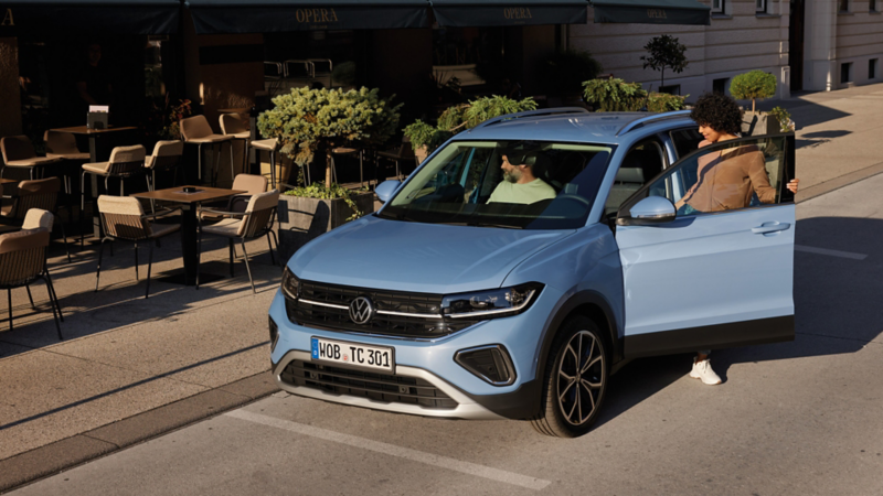 Eine Frau steigt auf der Fahrerseite in den blauen VW T-Cross, der vor einem urbanen Straßencafé parkt.
