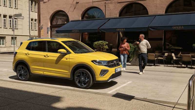  VW T-Cross in gelb parkt seitlich vor einem urbanen Straßencafé, ein Pärchen geht auf den Wagen zu.