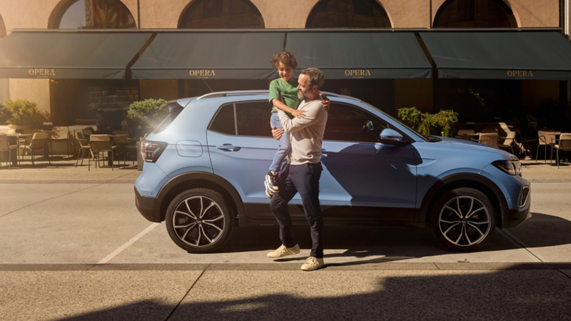 Ein Mann hält einen fröhlichen Jungen vor einem blauen VW T-Cross auf dem Arm.