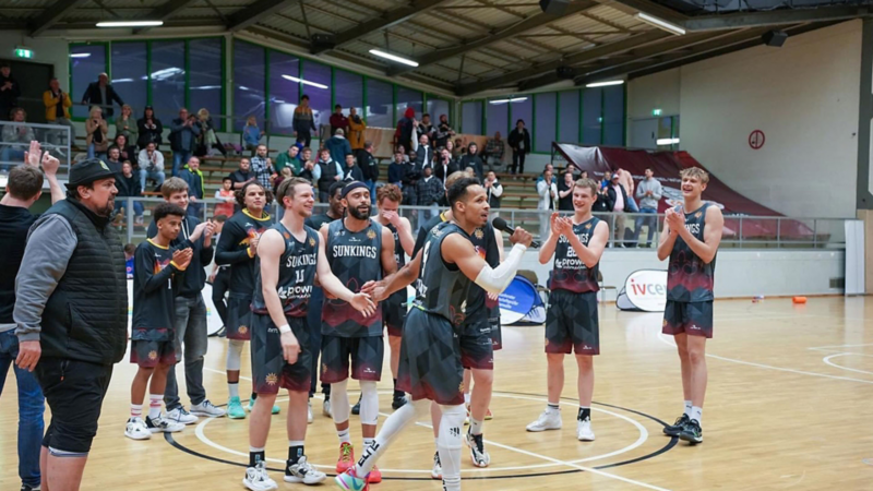 Basketballspieler in schwarzen Trikots mit 'SUNKINGS' feiern auf dem Spielfeld, Zuschauer im Hintergrund in der Halle