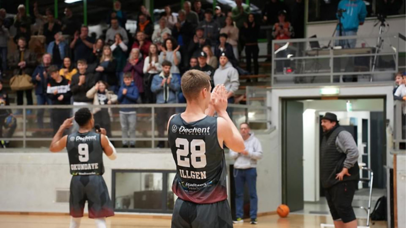 Zwei Basketballspieler in schwarzen Trikots mit den Nummern 9 und 28 klatschen vor Zuschauern in der Halle