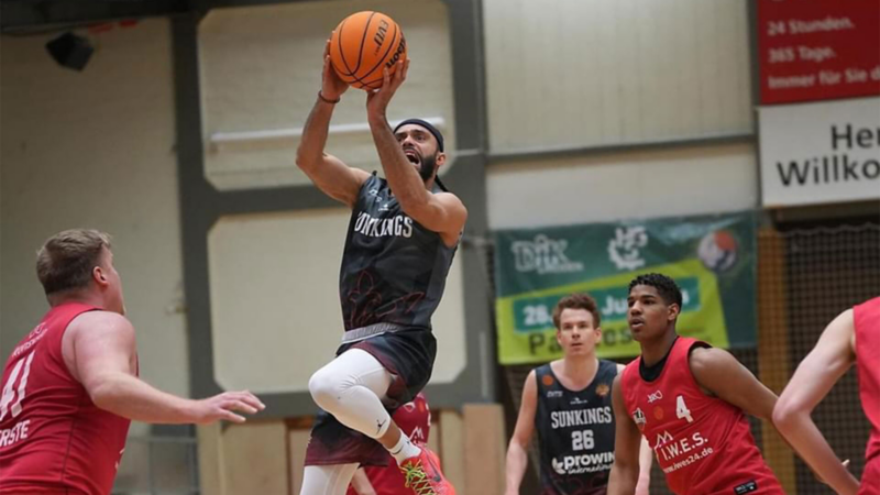 Basketballspieler in schwarzem Trikot springt zum Korbwurf, umgeben von Gegnern in roten Trikots in einer Halle