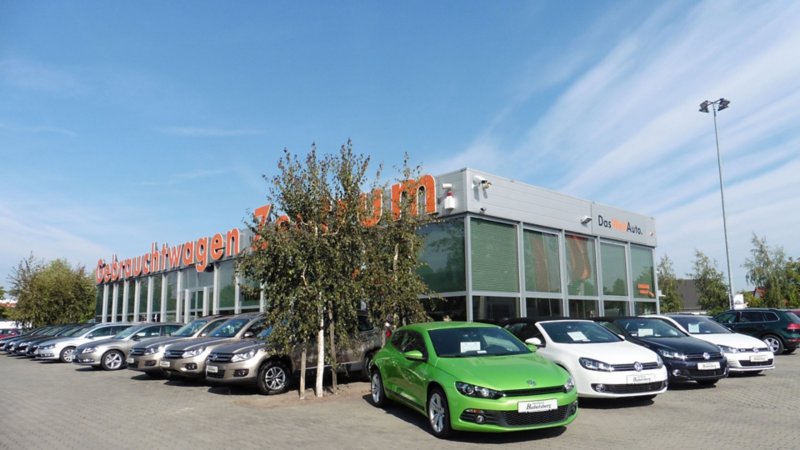 Autohaus mit mehreren geparkten Autos, darunter ein grüner und ein weißer Wagen im Vordergrund, bei klarem Himmel