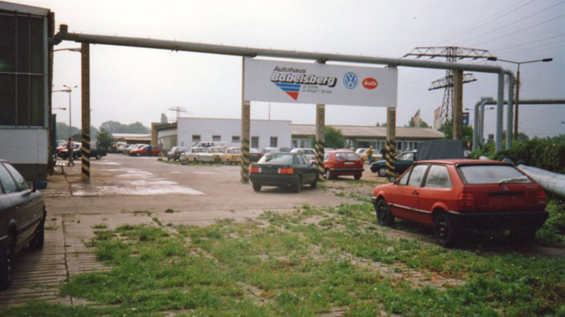 Außenbereich des Autohauses mit mehreren geparkten Autos, darunter ein rotes Fahrzeug im Vordergrund, und einem Schild mit den Logos von VW und Audi