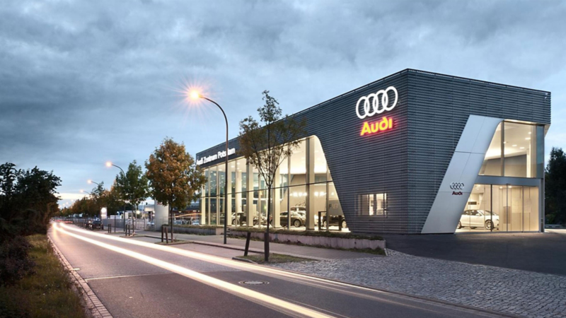 Audi Autohaus bei Dämmerung mit beleuchtetem Logo und modernen Glasfassaden an einer Straße mit Bäumen