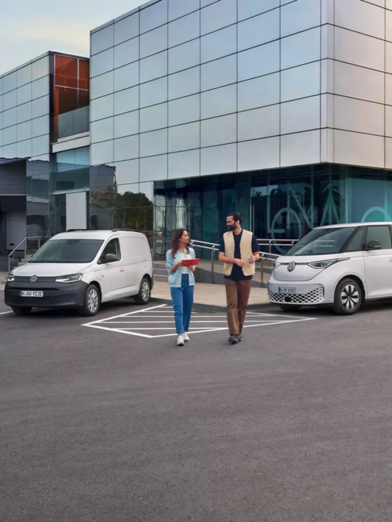 Ein Mann und eine Frau laufen auf einem Parkplatz auf dem Volkswagen Nutzfahrzeuge parken.