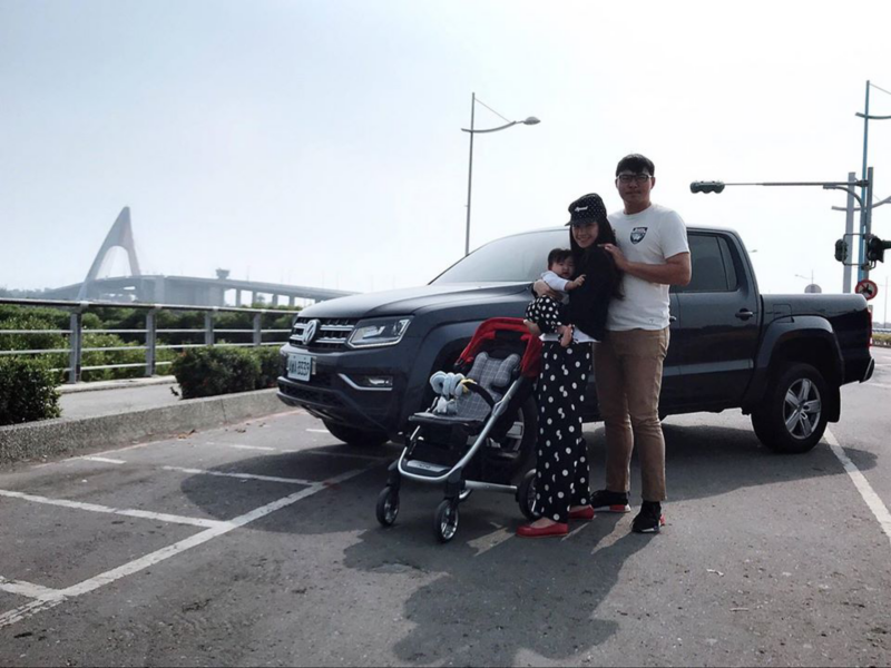 車主一家三口在珍珠黑色Amarok前方的合照，遠方可見造型大橋設計