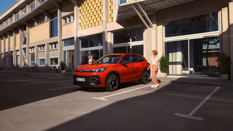 Na zdjęciu widać czerwonego Tiguana marki Volkswagen zaparkowanego na zewnętrznym parkingu przy nowoczesnym budynku, a obok niego stoi kobieta w jasnym stroju.